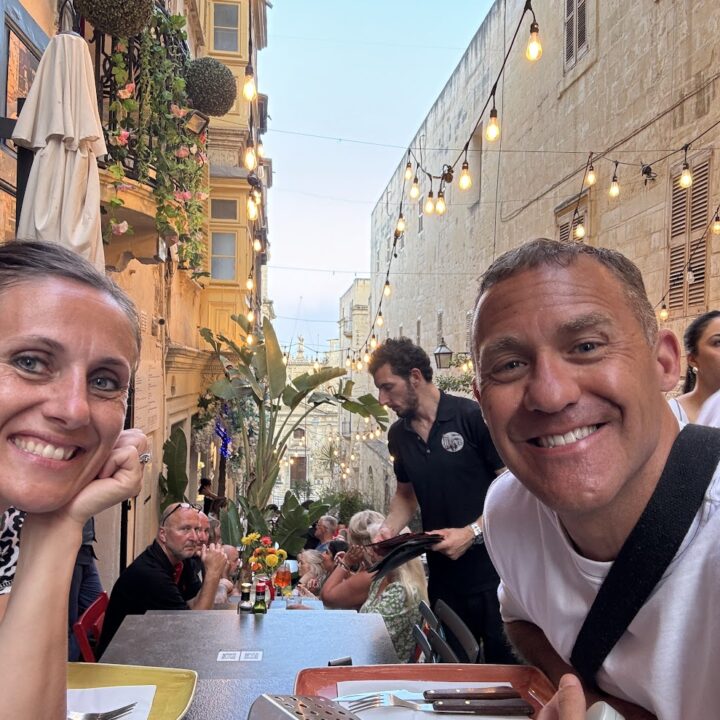 Paul and corene Hutchings sitting at a table with food and people around them in Malta