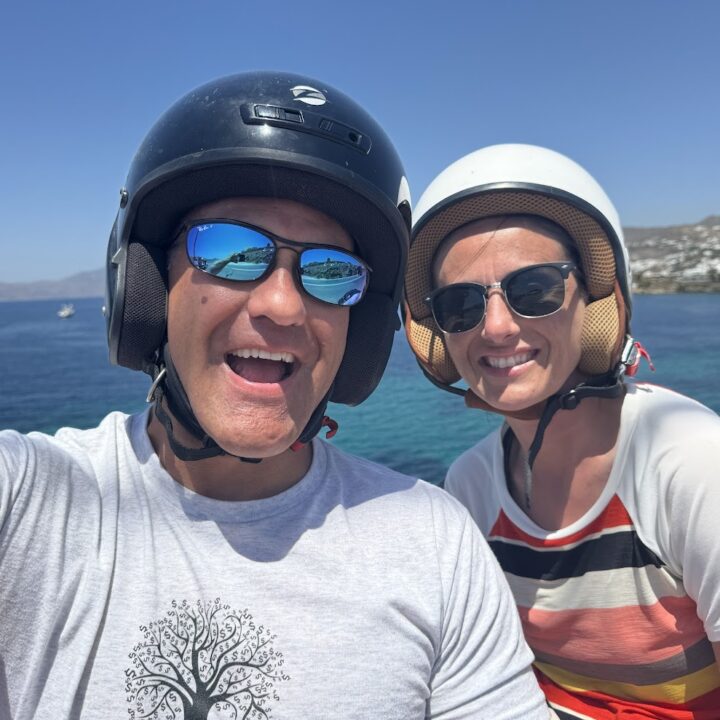 Paul and corene Hutchings wearing helmets and smiling on scooters in Mykonos